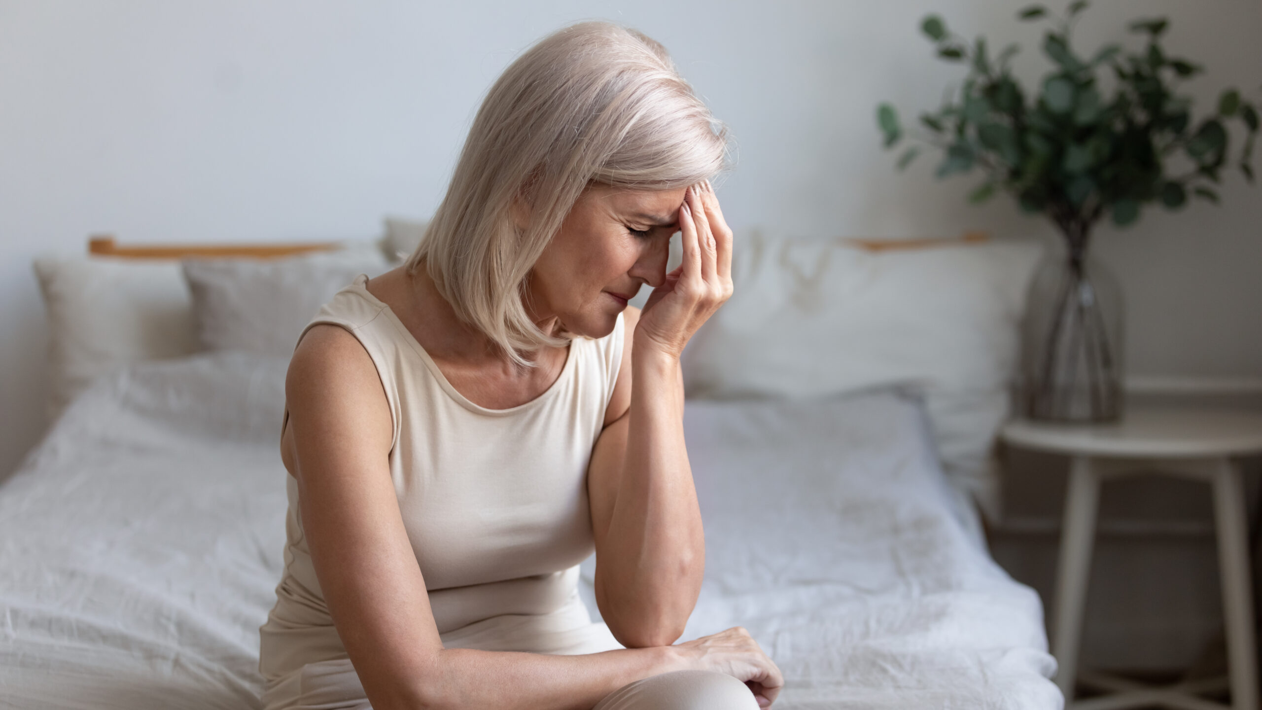 a senior woman sitting at the edge of her bed struggling with a headache and neck pain