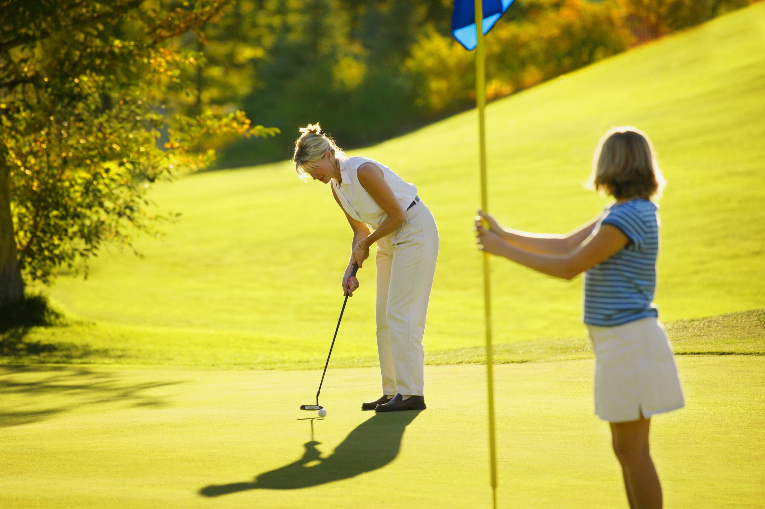 Two women out golfing on the green. One woman is putting, while the other is lifting the flag out of the pin.